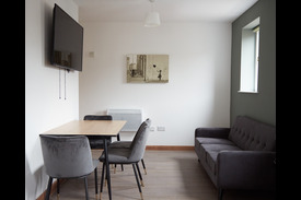 photograph of the communal area, a table with four chairs against the wall. A wall-mounted teleivison above. A three-seater sofa beneath a window.